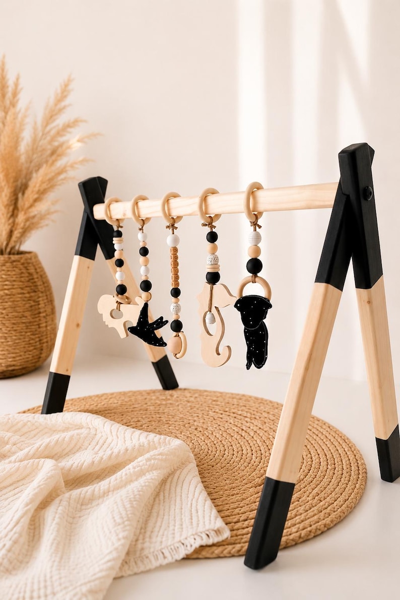 May include: A wooden baby play gym with hanging toys. The frame is natural wood with black accents. The hanging toys include wooden shapes, black and white beads, and wooden rings. The play gym is set on a woven mat.