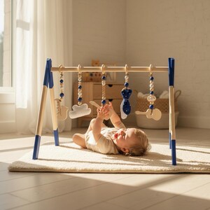 May include: A wooden baby play gym with blue accents and hanging toys. The toys include a cloud, a guitar, and a tie-shaped object. The baby is lying on a cream-colored rug, reaching for the toys. The gym is made of natural wood.