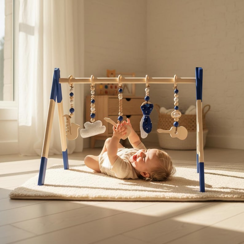 May include: A wooden baby play gym with blue accents and hanging toys. The toys include a cloud, a guitar, and a tie-shaped object. The baby is lying on a cream-colored rug, reaching for the toys. The gym is made of natural wood.