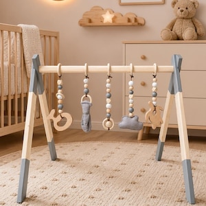 Peut inclure: Un portique d'éveil en bois avec une structure en bois clair et des pieds peints en gris. Cinq jouets suspendus sont fixés à la barre supérieure, comprenant des perles en bois, un nuage en tissu gris, une bande en tissu et des formes en bois.