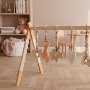 May include: Wooden baby play gym with hanging toys. The toys include a wooden heart, a wooden bear, a wooden turtle, and a peach-colored fabric shape. The gym has a natural wood finish with peach-colored accents on the legs.