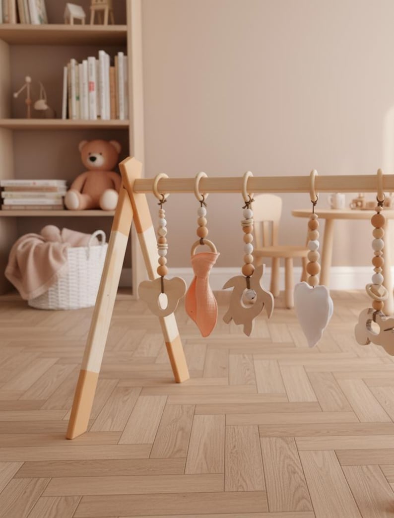 May include: Wooden baby play gym with hanging toys. The toys include a wooden heart, a wooden bear, a wooden turtle, and a peach-colored fabric shape. The gym has a natural wood finish with peach-colored accents on the legs.