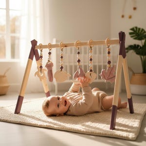 Könnte beinhalten: Baby-Spiel-Gymnastik aus Holz mit hängendem Spielzeug. Der Rahmen ist helles Holz mit dunkelvioletten Akzenten. Das hängende Spielzeug umfasst Holzformen, Stoffblätter und Perlen.