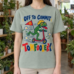 Pode incluir: Uma t-shirt verde sálvia com um gráfico de um sapo verde usando um capacete amarelo enquanto anda numa trotineta vermelha. O sapo segura uma bandeira vermelha com uma flor amarela. O texto diz OFF TO COMMIT TOMFOOLERY em letras coloridas.