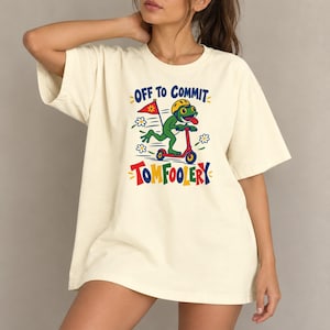 May include: A cream-coloured t-shirt featuring a graphic of a green frog wearing a yellow helmet while riding a red scooter. The frog holds a red flag, and the text reads "OFF TO COMMIT TOMFOOLERY" in blue and red letters with white daisy accents.