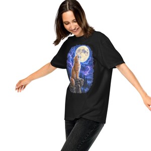 May include: A black short-sleeved t-shirt featuring a graphic print of an orange tabby cat sitting on a rocky cliff, howling at a large full moon in a dark blue sky filled with purple lightning bolts and silhouettes of pine trees.