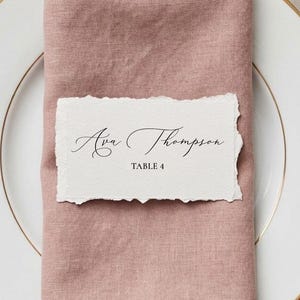 May include: A white dinner plate with a gold rim, topped with a dusty rose linen napkin and a rectangular place card featuring torn edges. The card displays the text Ava Thompson and TABLE 4 in black ink, accompanied by a silver fork, knife, and spoon.