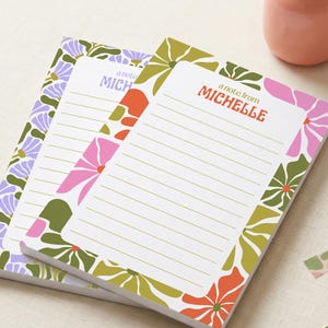 May include: Three floral-patterned notepads with lined pages. The top notepad features the text "a note from MICHELLE" in orange. The floral designs include pink, green, and orange flowers. A pink vase with flowers, a pen, and decorative tape are also visible.