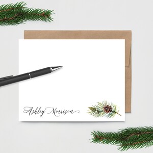 May include: A white card with a brown envelope. The card has a watercolor illustration of a pine branch with a pine cone. The text "Ashley Morrison" is written in cursive at the bottom of the card.