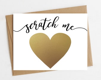 Puede incluir: Una tarjeta blanca con el texto "scratch me" en escritura cursiva negra. Un corazón de lámina dorada está centrado en la tarjeta.