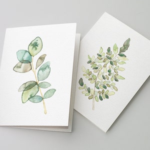 May include: Two watercolor greeting cards with green foliage designs. The card on the left features a branch of eucalyptus leaves in shades of green and gray. The card on the right features a fern with green leaves.