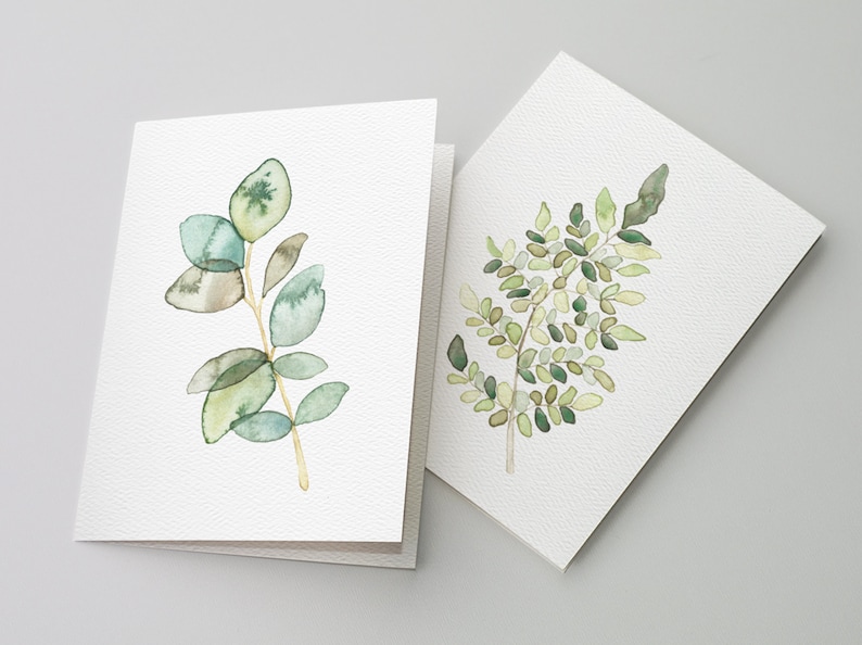 May include: Two watercolor greeting cards with green foliage designs. The card on the left features a branch of eucalyptus leaves in shades of green and gray. The card on the right features a fern with green leaves.