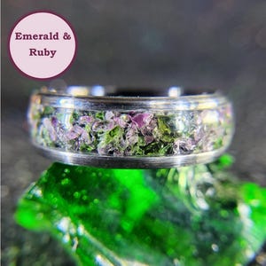 May include: A silver ring with a band of crushed emerald and ruby gemstones. The ring is set on a green background.