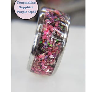 May include: A silver ring with a band of pink, purple, and black glitter embedded in clear resin. The ring is labeled "Tourmaline Sapphire Purple Opal".