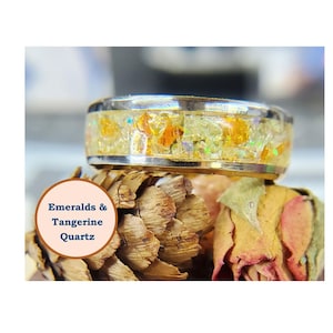 May include: A gold ring with a band of clear resin embedded with small pieces of green and orange gemstones. The text "Emeralds & Tangerine Quartz" is visible in the image.
