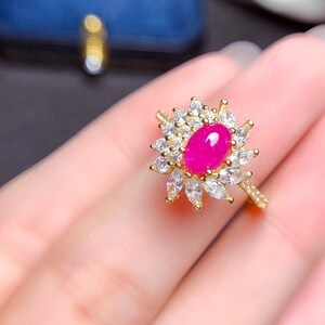 May include: A gold ring featuring a vibrant fuchsia oval gemstone, surrounded by a halo of clear, sparkling accent stones. The ring's design resembles a flower, with the central stone as the focal point. The band is also gold.