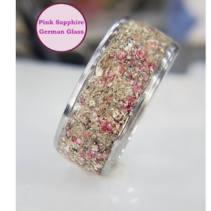May include: A silver ring with a band of pink and gold crushed glass. The ring is labeled "Pink Sapphire German Glass".