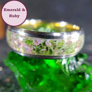 May include: A gold and silver ring with a band of embedded green and pink gemstones. The ring is set on a green surface.