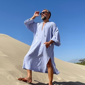 May include: A light blue linen tunic with long sleeves and a split hem. The tunic is worn with brown leather sandals. The person is standing on a sand dune under a clear blue sky.