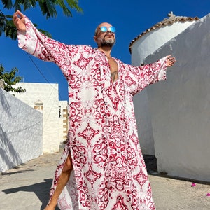 May include: A person wearing a long, white and pink patterned robe with a deep V-neckline. The robe has a geometric design and is open in the front. The person is standing with their arms outstretched, wearing sunglasses.