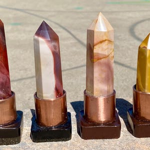 May include: Four polished gemstone obelisks in shades of red, cream, and yellow. Each obelisk is set in a copper-colored metal sleeve and mounted on a dark square base. The obelisks vary in color and pattern.