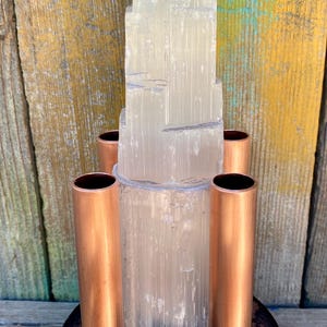 May include: A vertical, translucent white selenite crystal tower stands centered on a circular, dark metal base. Four smooth, cylindrical copper pipes are arranged symmetrically around the base of the crystal, creating a structured, multi-material display piece.