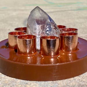 May include: A brown, circular crystal holder with six copper-colored metal cylinders arranged around a central, clear quartz crystal point. The holder is designed to display crystals and gemstones.