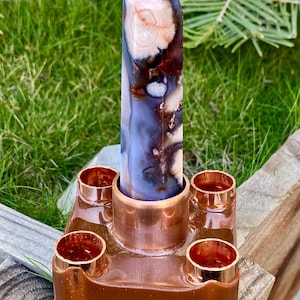 May include: A tall, polished crystal obelisk with a pointed top, featuring a blend of white, brown, and blue hues. It is set in a copper-colored metal base with four cylindrical holders. The background shows green grass and foliage.
