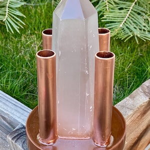 May include: A clear quartz crystal point stands in the center of a copper-colored metal base with four cylindrical holders. The base is round and sits on a wooden surface, with green grass and pine needles in the background.