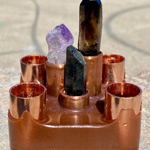 May include: A copper-colored crystal holder with five cylindrical openings. The holder contains a variety of crystals, including a large, dark smoky quartz, a purple amethyst, and a black tourmaline. The crystals are arranged in the center and around the edges of the holder.