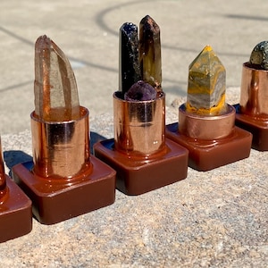 May include: A row of six decorative crystal and gemstone pieces. Each piece features a different crystal or gemstone set in a copper-colored metal casing atop a brown square base. The crystals and gemstones include clear, black, yellow, and green varieties.