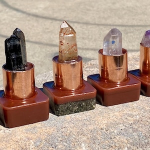 May include: Six crystal lipstick holders with copper-colored metal tubes and brown bases. The crystals vary in color, including smoky quartz, black tourmaline, clear quartz, and amethyst. The holders are arranged in a row on a stone surface.