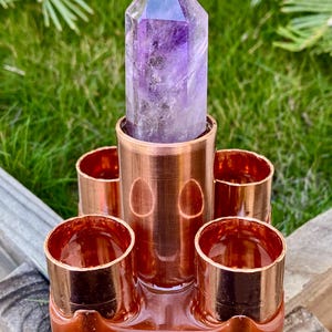 May include: A copper-coloured crystal holder with a large, pointed amethyst crystal. The holder has four small cup-like openings around a central, larger opening. The background is green grass and wood.