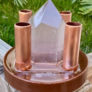 May include: A brown, circular base holds four copper tubes and a clear, pyramid-shaped crystal. The crystal is centered, with the tubes evenly spaced around it. The base has a shallow, liquid-filled basin.