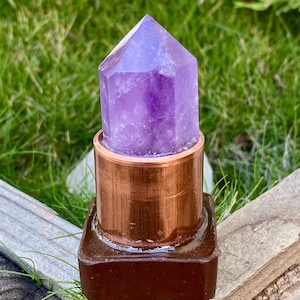 May include: A purple amethyst crystal point sits atop a copper cylinder and a dark brown square base. The crystal is a vibrant purple color and has a pointed top. The copper cylinder has a polished finish. The base is a dark brown color.
