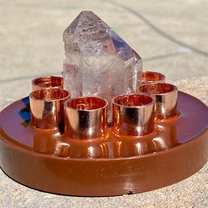 May include: A brown, circular candle holder with six copper-colored candle cups surrounding a large, clear crystal. The crystal is a light purple color and is the focal point of the piece. The candle holder is made of a shiny, smooth material.