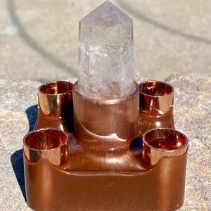 May include: A copper-colored crystal holder with a clear quartz crystal. The holder has a square base with four cylindrical cup-like structures around a central, larger cylinder that holds the crystal. The crystal is a clear, pointed prism.