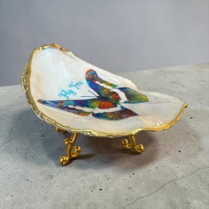 May include: A white oyster shell dish with a gold-painted rim and three gold-toned metal legs. The interior features a colorful butterfly graphic with the text Fly Free in blue script. The shell has a smooth, pearlescent finish and curved edges.