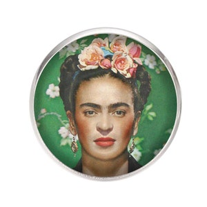 Può includere: Un bottone rotondo color argento con un'immagine colorata di Frida Kahlo. Indossa una corona di fiori e ha un'espressione seria sul viso. Lo sfondo è un motivo floreale verde.