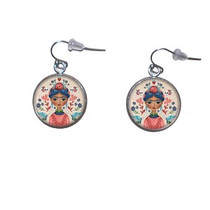 Könnte beinhalten: Ein Paar silberne Ohrringe mit einem runden, cremefarbenen Hintergrund, auf dem eine bunte Illustration einer Frau mit einer Blumenkrone und blauen Haaren zu sehen ist. Die Frau trägt ein rosa Kleid und eine blaue Halskette. Die Ohrringe haben einen silbernen Haken.