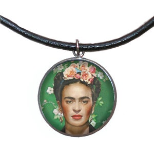 Può includere: Una collana in pelle nera con un ciondolo rotondo in argento che presenta un ritratto di Frida Kahlo con fiori nei capelli. Lo sfondo è verde con fiori bianchi.