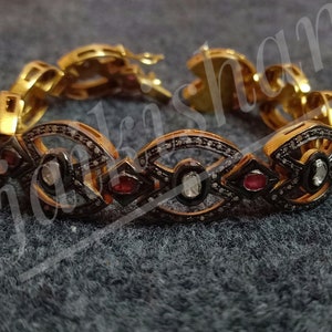 May include: A gold bracelet with a black oxidized finish. The bracelet features a repeating pattern of oval-shaped settings with white diamonds and red rubies.