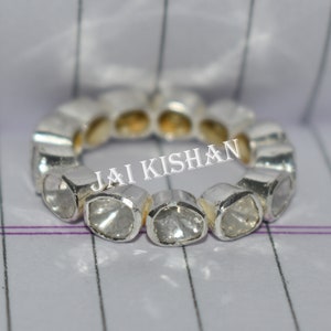 May include: A silver ring featuring a row of round, clear gemstones. The ring is displayed on a white and grey striped surface. The text "JAI KISHAN" is visible on the ring.
