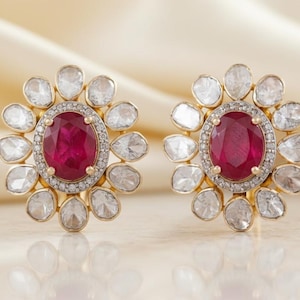 Puede incluir: Un par de pendientes de oro. Cada pendiente presenta una gran gema ovalada de color rojo intenso rodeada por un halo de pequeñas gemas transparentes. El borde exterior de cada pendiente tiene un diseño floral con gemas transparentes.