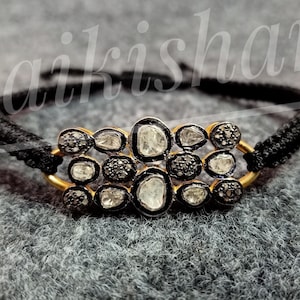 May include: A black cord bracelet with a gold setting featuring a cluster of white and black diamonds.