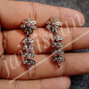 以下が含まれることがあります： 花柄のシルバー製ドロップイヤリングのペア。各イヤリングは、中央にピンクの石がついた小さな花と、ピンクの石がついた葉の垂れ下がったチェーンが特徴です。