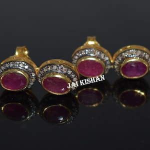May include: Close-up of four gold-toned earrings with oval, deep red gemstones. Each earring is surrounded by a ring of small, clear stones and a dark metal border. The earrings are displayed on a reflective black surface.