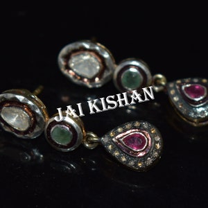 May include: A pair of silver earrings with a teardrop-shaped pendant featuring a red gemstone surrounded by smaller diamonds. The earrings also have a smaller round pendant with a green gemstone and a larger round pendant with a white gemstone.