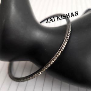May include: A silver bangle bracelet with a row of small, clear stones around the edge. The bracelet is on a black surface and the text "JAI KISHAN" is visible in the background.
