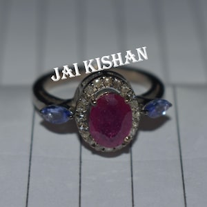 Puede incluir: Un anillo de plata con un gran rubí ovalado rodeado de diamantes más pequeños y dos piedras azules más pequeñas a cada lado. El anillo está inscrito con el texto "JAI KISHAN".